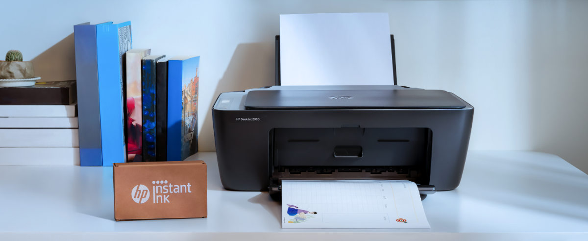 Black HP DeskJet 2955 printer on a white desk actively printing a document, with an HP Instant Ink box in the foreground and books in the background.