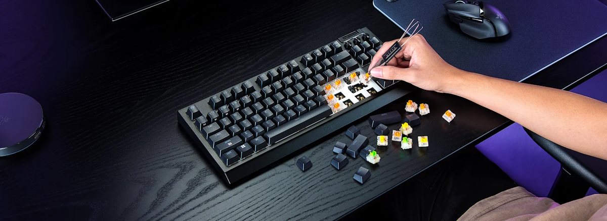 A person repairs a mechanical keyboard, using tweezers, with several colorful key switches and keycaps scattered on a black desk.