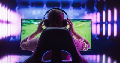 A person viewed from behind, wearing large over-ear headphones and adjusting them with both hands. The person is seated in a dark gaming chair with subtle blue glowing accents, facing a large monitor displaying a bright green video game scene. The dark background is illuminated by vibrant vertical purple and blue neon light strips, creating an immersive and focused gaming environment.