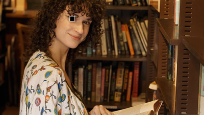 A woman in a library looking towards the camera - a small white square over one of her eyes represents Eye AF for video