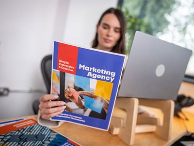 A lady holding a colour marketing agency document while sat at a desk in a home office environment