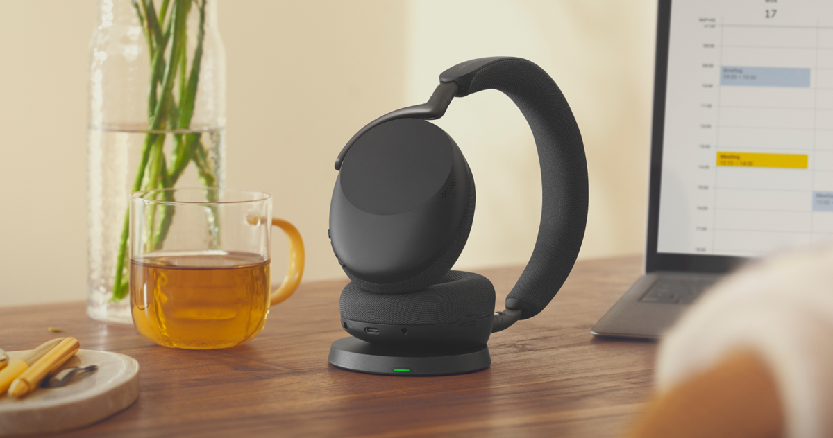 Black Jabra Evolve3 headset on its charging stand next to a cup of tea and an open laptop.