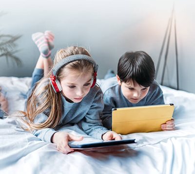 Two children lying on a bed using their tablets.