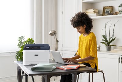 Female with yellow shirt working in home office with a laptop and printer at her desk.