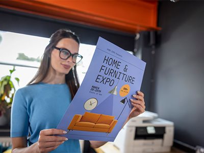 A lady holding a colour home and furniture expo poster with a Brother MFC-J6960DW in the background
