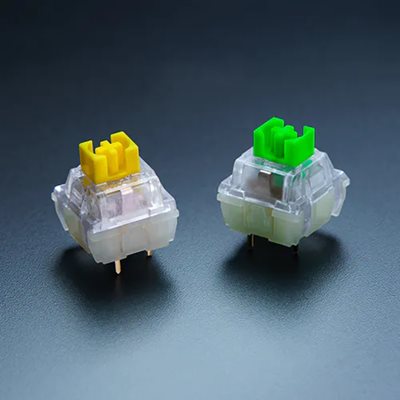 Two transparent mechanical keyboard switches with yellow and green stems on a dark surface, showing internal contact mechanisms.