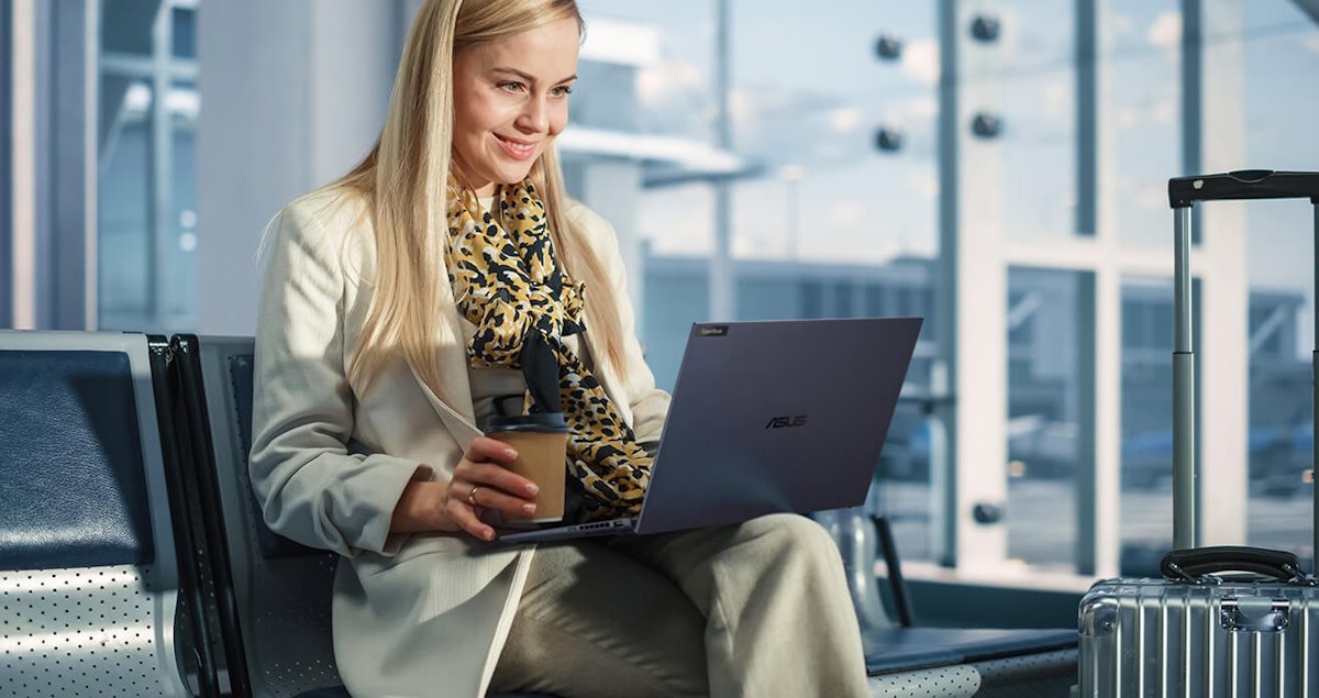 A business traveler working on a laptop at the airport, holding a coffee and sitting next to her luggage.