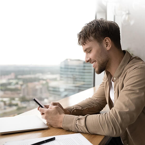 Ein Mann sitzt in einem Café am Fenster, lächelt und schaut auf sein Smartphone.