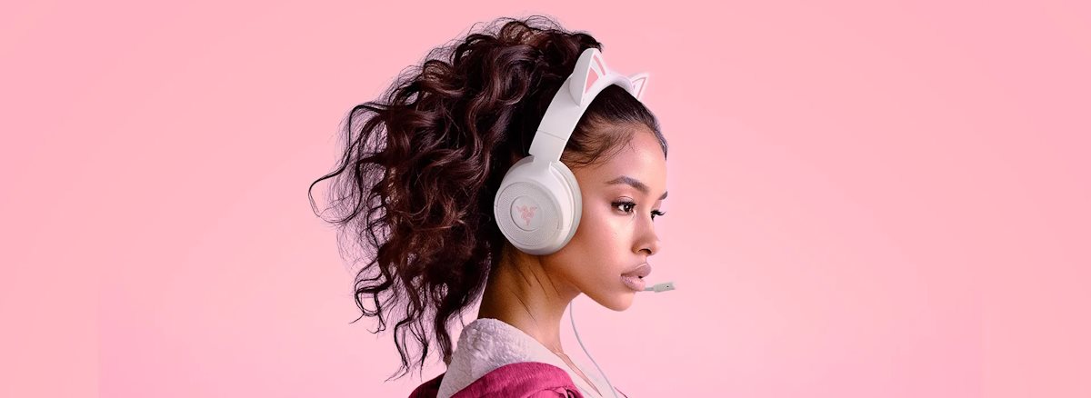 Young woman with curly hair wearing pink and white bunny-eared headphones with a microphone, against pink background.