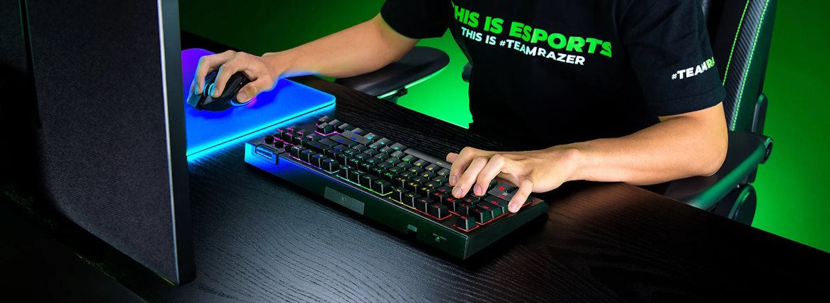 Person using a gaming keyboard, mouse, and monitor in a dark setting with green and RGB lighting; wearing a black T-shirt with green and white text.