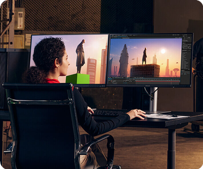 A woman works with digital 3D modeling on dual monitors, featuring a futuristic cityscape and statue. The setting conveys creativity and focus.