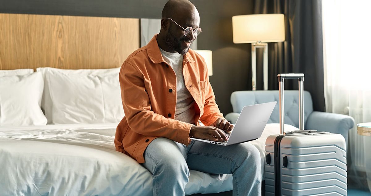 A business traveler working remotely on a laptop in a hotel room, with a suitcase nearby.
