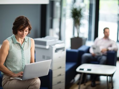 A woman using a notebook computer while sat next to a multifunction printer in an SMB environment
