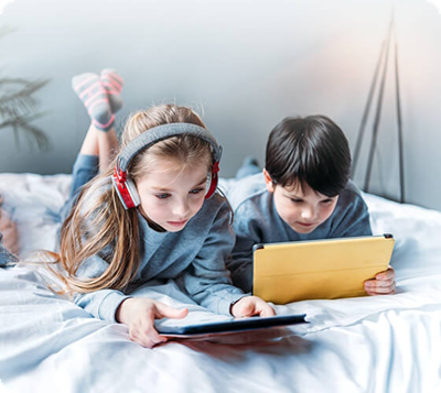 Two children lying on a bed using their tablets.