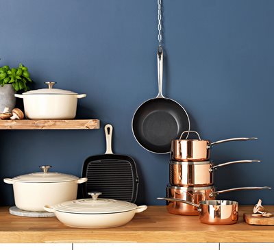 Cast iron casserole dishes and a copper triply pan set displayed on a wooden worktop against a blue wall.