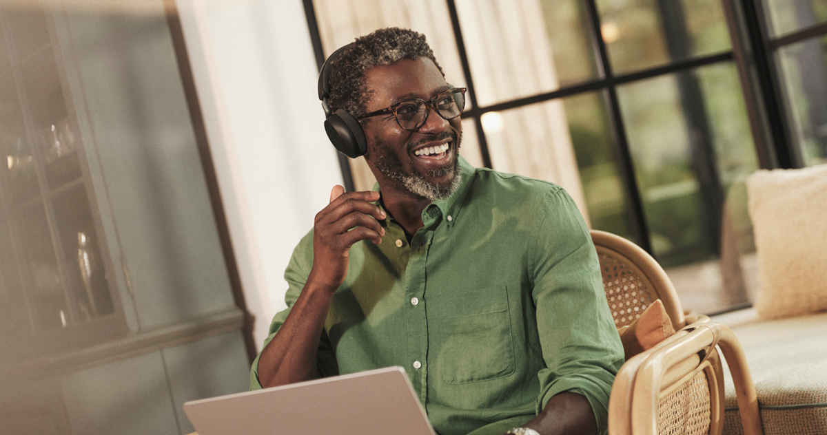Person i grøn skjorte med et Jabra Evolve3 75-headset, der rører ved ørekoppen, mens vedkommende er i et opkald derhjemme med en bærbar computer foran sig.