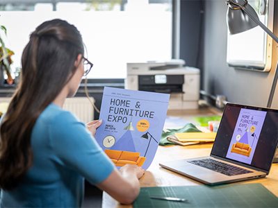 A lady holding a colour home and furniture expo poster while sat at a desk in a home office environment