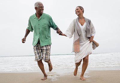 Một cặp vợ chồng trung niên/đã nghỉ hưu, khỏe mạnh, nắm tay nhau chạy trên bãi biển gần mặt nước.