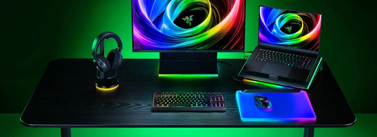A gaming setup with a large Razer monitor, RGB mechanical keyboard, RGB mouse on a mouse pad, a laptop with a colorful display, and black headphones.