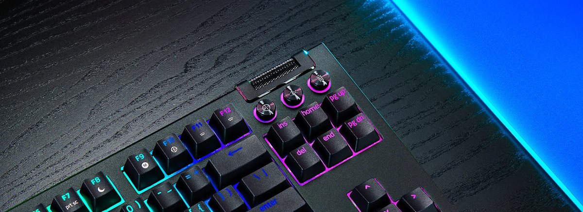 RGB backlit gaming keyboard with a metal scroll wheel and keys labeled F7 to F12, on a dark textured desk with blue LED edge lighting.