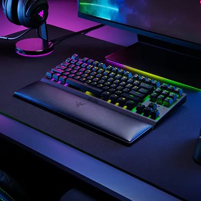 RGB backlit mechanical keyboard with wrist rest on a dark gaming desk, monitor, and headphones with purple lighting.