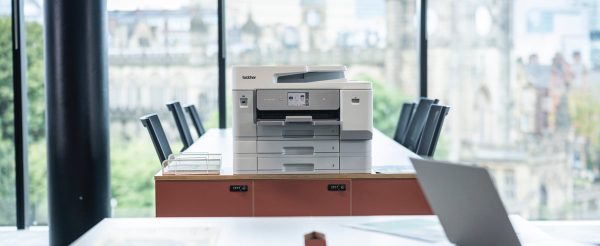 A Brother MFC-J6977DW on a long desk in front of a glass wall in an office environment